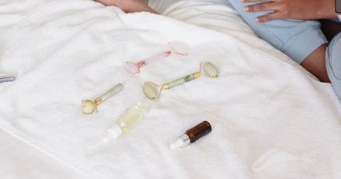 African american woman arranging jade and rose quartz facial rollers with serums on bed