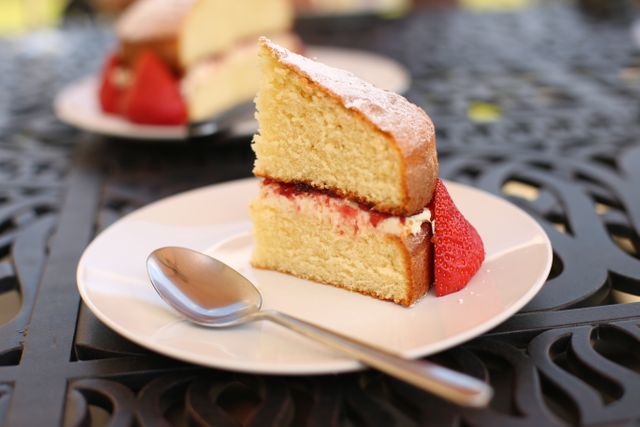 This delectable slice of Victoria sponge cake features layers of fluffy cake filled with cream and jam, garnished with a fresh strawberry, and sits on a white plate with a spoon beside it. Ideal for use in food blogs, dessert menus, culinary magazines, and social media posts showcasing homemade treats and recipes.