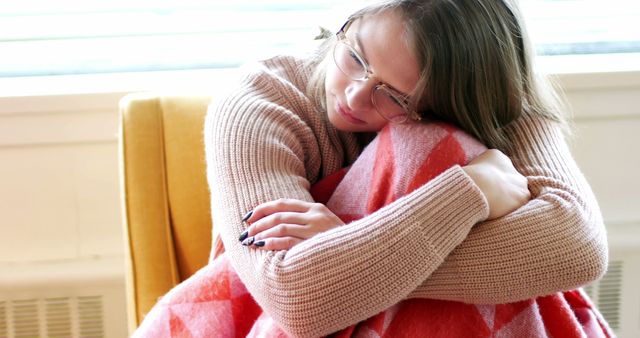 Young woman sitting in cozy chair with red patterned blanket, showcasing a peaceful and introspective moment. She is wearing a warm sweater and glasses, looking thoughtful and relaxed. Perfect for use in lifestyle blogs, mental health articles, and advertising materials focused on comfort and relaxation.