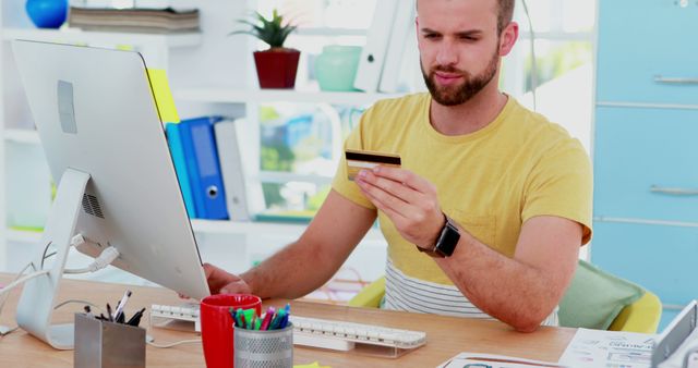 Young man in casual clothes online shopping with credit card at home office desk. Scene includes computer, stationery, files, and modern decor. Useful for depicting e-commerce, technology, remote work, and financial transactions. Ideal for websites, blogs, or articles about online banking, digital payments, or remote working environments.