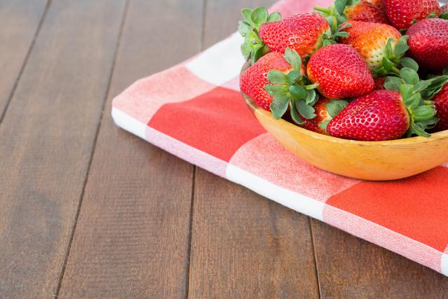 Bowl of juicy, ripe strawberries placed on a red and white checkered napkin on a wooden surface. Ideal for use in content related to healthy eating, summer snacks, fresh produce, or rustic kitchen settings.