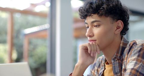 Young man working remotely at laptop by glass doors with natural light and patio view