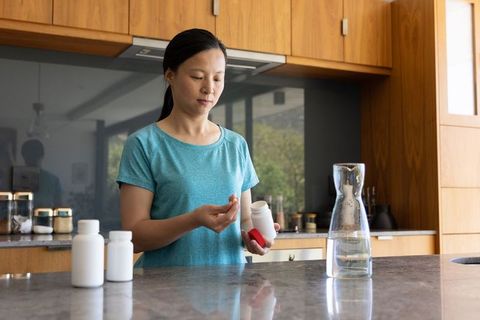 Mature asian woman taking daily supplements in modern kitchen for wellness routine and hydration