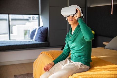 Young man wearing vr headset on sunlit bed in modern bedroom exploring virtual reality