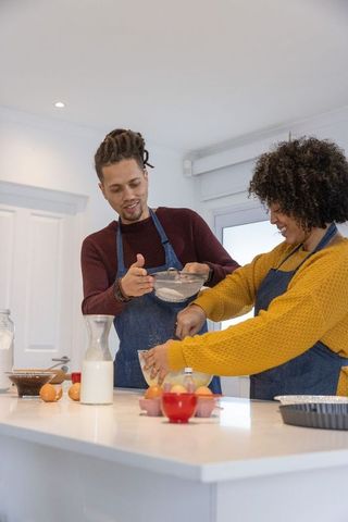 African american couple baking together at kitchen island, sifting flour and cracking eggs