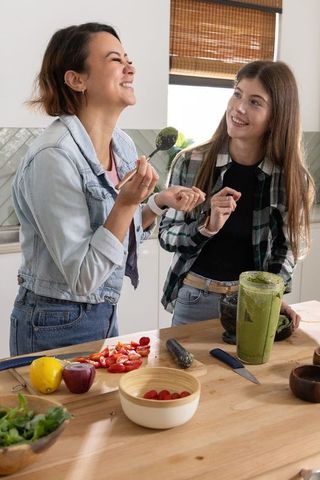 Mother and teen daughter laughing while tasting green smoothie at kitchen counter