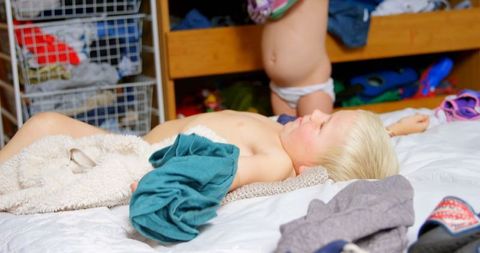 Young Child Relaxing on Bed with Sibling Dressing Nearby