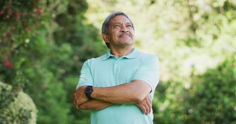 Senior man standing folding arms gazing upward in sunlit garden wearing mint polo, watch