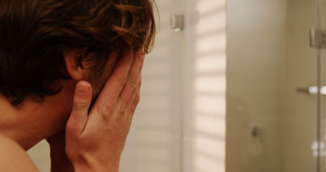 The man in the bathroom is covering his face with his hands while standing in front of mirror. He seems to be in an emotional or distressed state. This image can be used in articles and blogs about mental health, personal reflection, dealing with stress, feelings of being overwhelmed, or emotional challenges.