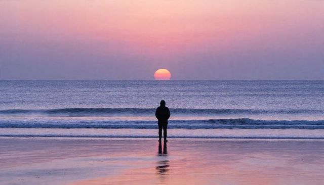 Solitary silhouette standing on wet sand watching pastel sunrise reflecting on calm ocean