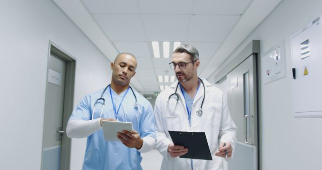 Two doctors walking through hospital corridor, discussing medical patient data. One doctor holding a tablet, another holding a clipboard. Suitable for use in healthcare, teamwork, patient care, medical communication, and hospital-related themes.