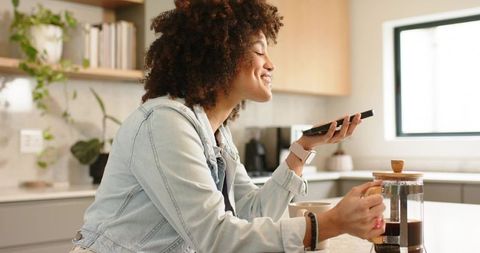 Young woman enjoying coffee and voice messaging on smartphone at bright kitchen counter
