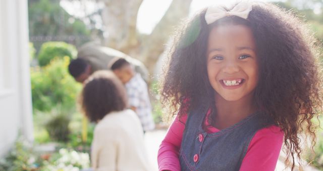 A young African American girl with curly hair is smiling at the camera, while her family is seen in the background enjoying time together outdoors. This image is perfect for use in advertising materials or blog posts focused on family activities, childhood happiness, or outdoor play. It can also be used in educational content promoting family values and quality time.