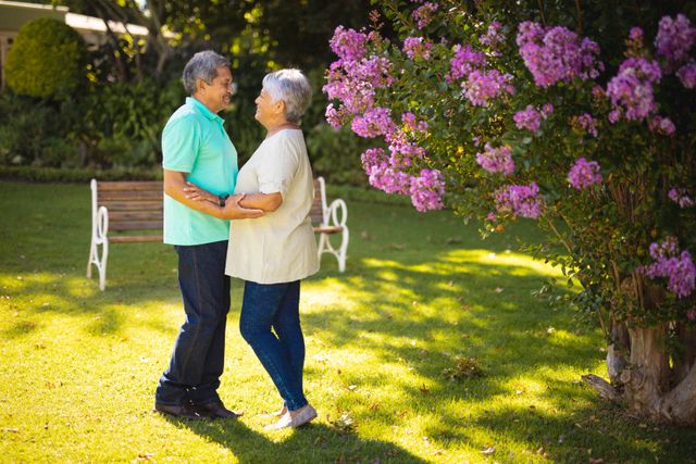 Senior couple standing close, holding hands and looking at each other lovingly in a park with blooming flowers. Ideal for use in content related to senior lifestyle, love in later years, retirement, and outdoor activities for elderly couples.
