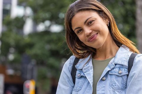 Young woman smiling and tilting head on city sidewalk wearing denim jacket and backpack