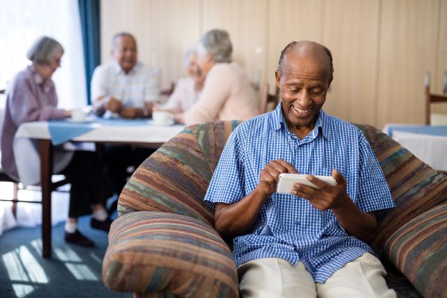 Senior man sitting on sofa using smartphone while friends socialize in background. Ideal for illustrating technology use among elderly, senior living communities, and social interactions in retirement homes.
