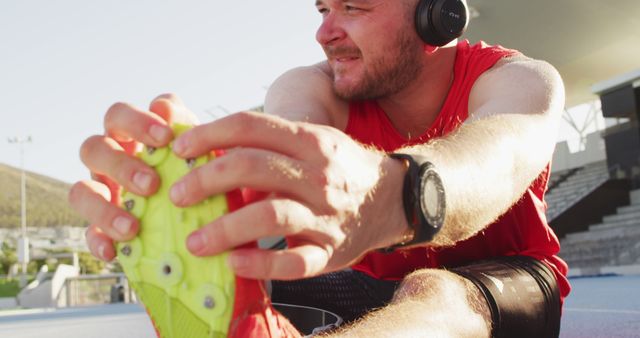 Male athlete wearing red shirt stretching on outdoor track while listening to music on headphones. Ideal for use in sports, fitness, and health-related materials, emphasizing preparation, warm-up, or running.