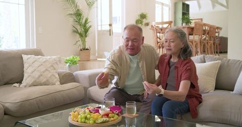 Older asian couple reaching for healthy fruit platter with nutritional labels in bright living room