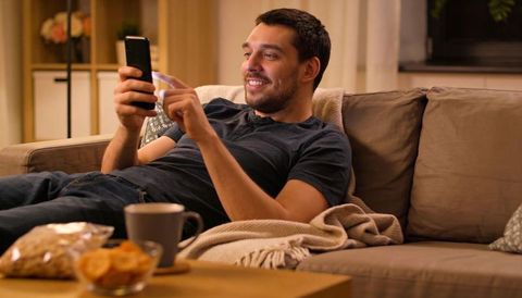 Young man relaxing on sofa scrolling smartphone smiling cozy living room evening lifestyle