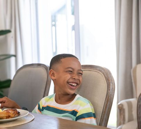 Laughing african american boy sitting at dining table enjoying meal in bright home