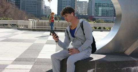 Young man sitting on urban bench wearing gray hoodie and earbuds holding phone and coffee