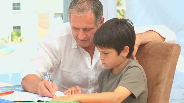 Grandfather helping grandson with homework at living room table. They are showcasing family bonding through education. Grandfather is guiding the child who is focused on writing. Ideal for materials related to family relationships, education, intergenerational bonding, and at-home learning activities.