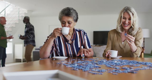 African american and caucasian senior women sitting by table doing puzzles drinking tea. senior retirement lifestyle friends socialising.
