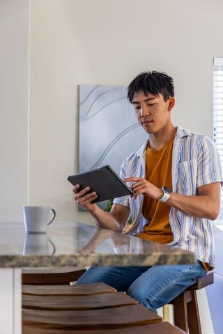 Asian man reading tablet at kitchen bar with coffee mug, smartwatch and natural light