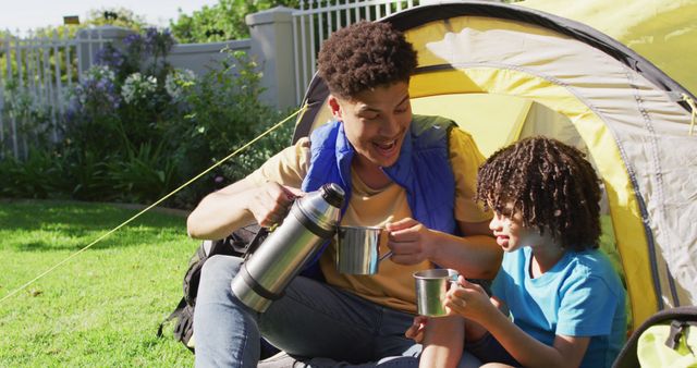 Father and son sitting by tent sharing hot drink from thermos. Ideal for portraying family bonding, camping trips, and outdoor adventure.