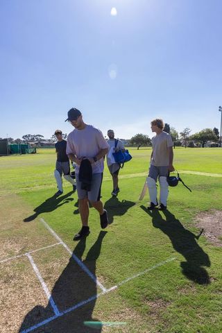 Diverse male cricket teammates walking across sunlit pitch carrying bats and batting pads
