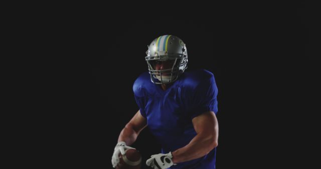 Football player wearing a blue jersey and full protective gear holding a football, captured in action against a dark background. Can be used for sports promotions, athletic training materials, or motivational posters.