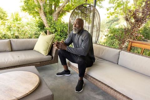 Senior african man sitting on patio sofa with rattan swing, enjoying quiet garden lounge