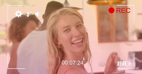 Smiling blonde woman at kitchen bar holding drink with straw during friends gathering