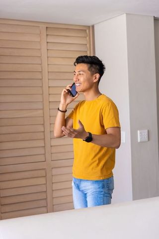 Young asian man talking on smartphone at home wearing mustard t-shirt and smartwatch