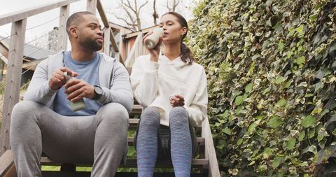 Diverse couple relaxing on outdoor steps drinking from reusable bottles in cozy activewear