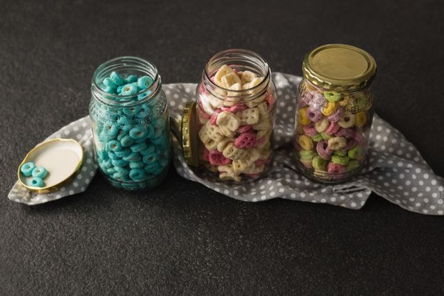 Three glass jars filled with different colorful breakfast cereals are placed on a dark background. This image can be used for promoting healthy breakfast options, food storage solutions, or kitchen organization tips. It is ideal for blogs, websites, and advertisements related to nutrition, diet, and healthy living.