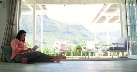 Mature asian woman relaxing and reading paperback by open terrace with mountain view
