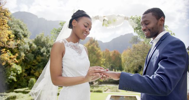 Couple exchanging rings during their wedding ceremony outdoors. Perfect for use in wedding planning materials, marriage counseling brochures, and romantic greeting cards.
