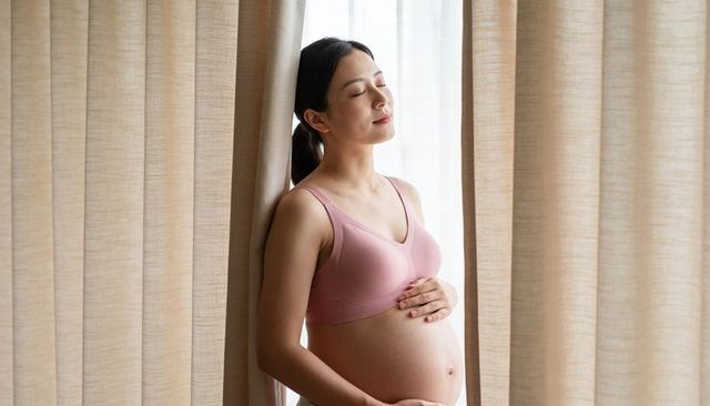 Pregnant woman standing at window cupping baby bump in soft natural light