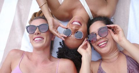 Laughing mixed-ethnicity friends lying on striped towel wearing round sunglasses under sun
