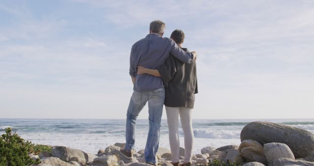 Mature couple standing on a rocky beach, embracing while looking at the ocean. Ideal for themes of love, companionship, togetherness, retirement, relaxation, vacation, and scenic oceanside enjoyment. Perfect for advertisements, articles, blog posts on relationships or travel.