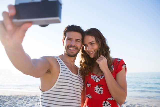 Young couple enjoying a sunny day at the beach, taking a selfie to capture the moment. Ideal for use in travel brochures, vacation advertisements, relationship blogs, and social media campaigns promoting outdoor activities and summer fun.