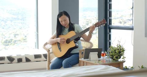 Asian woman playing acoustic guitar in sunlit bedroom corner, practicing music at home