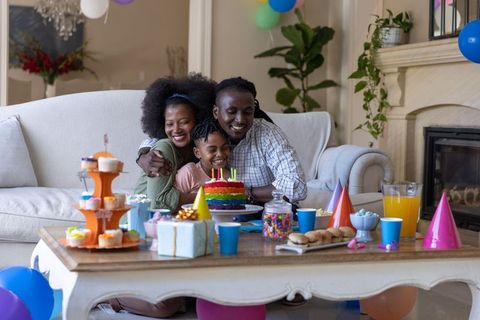 African american family celebrating birthday hugging and blowing candles on rainbow cake