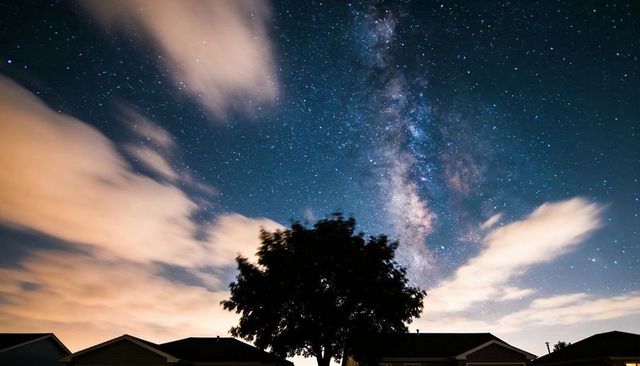 Milky way rising over suburban rooftops with silhouetted tree and glowing clouds at night