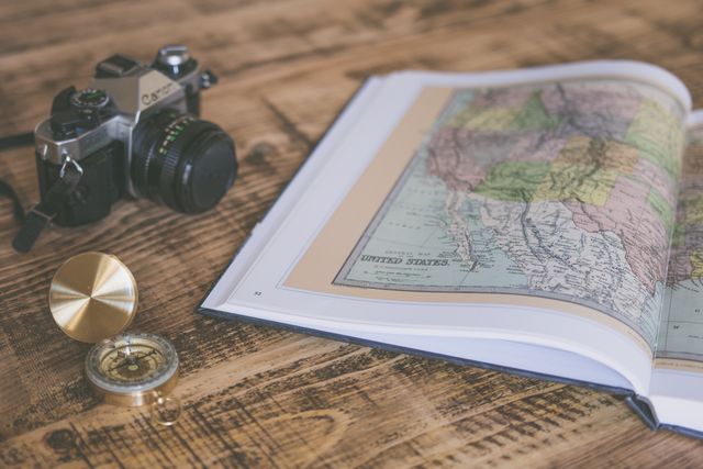 This close-up image showcases a vintage film camera, an open book featuring a map, and a brass compass on a rustic wooden table. It evokes a sense of nostalgia, travel, and adventure. Perfect for use in travel blogs, adventure gear promotions, and vacation planning websites.