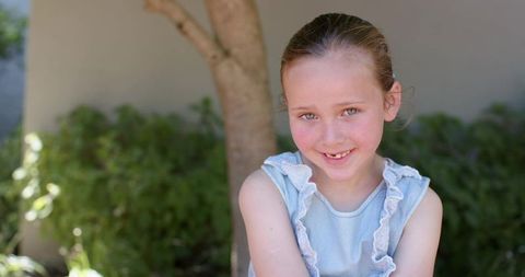 Sunlit young girl smiling in backyard garden wearing light blue ruffled dress, natural portrait