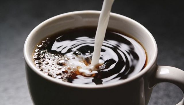 Pouring milk into black coffee creating creamy swirl, bubbles and ripples in white mug