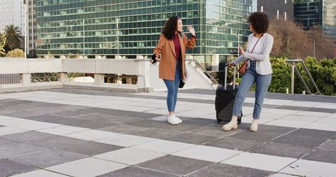 Two stylish women waving on urban rooftop terrace with rolling suitcase and smartphones