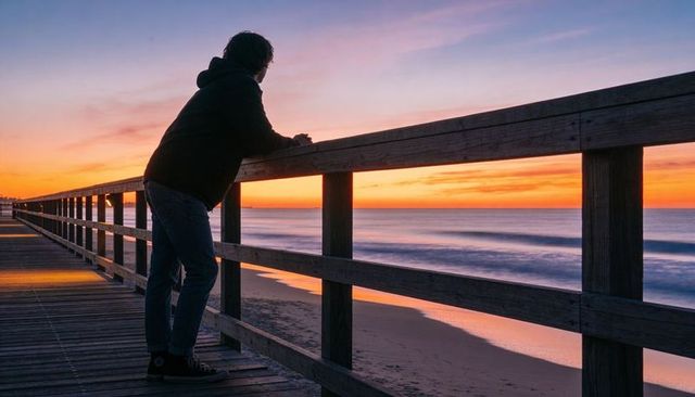 Man leaning on pier railing watching ocean sunset silhouette at twilight